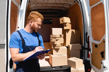 Dışarıdaki paketlerle kamyonun yanında pano tutan tulumlu bir yükleyici. 