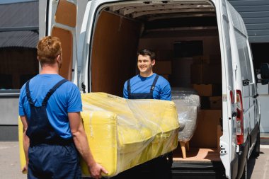 Selective focus of movers holding sofa in stretch wrap near truck on urban street  clipart