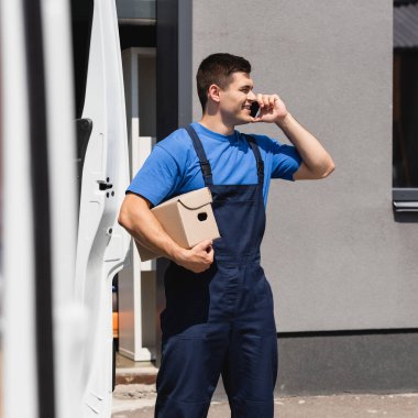 Akıllı telefondan konuşan ve şehir caddesinde kamyonun yanında karton kutu tutan yükleyicinin seçici odak noktası 