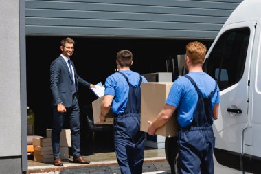 Selective focus of businessman holding clipboard while movers carrying packages near truck and warehouse outdoors clipart