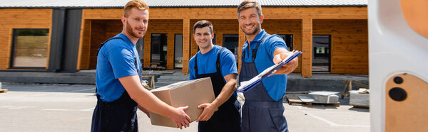 Горизонтальный урожай перевозчиков с планшетом и картонной коробкой, смотрящих на камеру возле двери грузовика на городской улице 