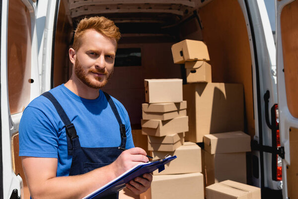 Selective focus of loader in uniform holding clipboard and pen with cardboard boxes in truck at background 