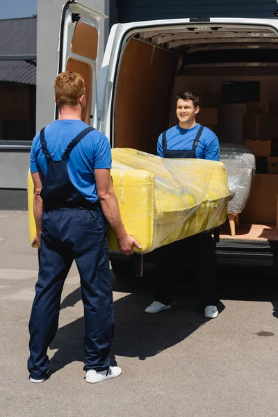 Selective focus of loader carrying couch in stretch wrap with colleague near truck on urban street