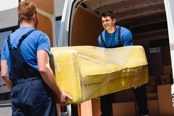 Selective focus of movers unloading sofa in stretch wrap in truck outdoors 