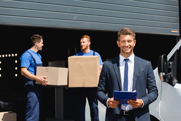 Selective focus of businessman holding clipboard while movers holding carton boxes near truck and warehouse 