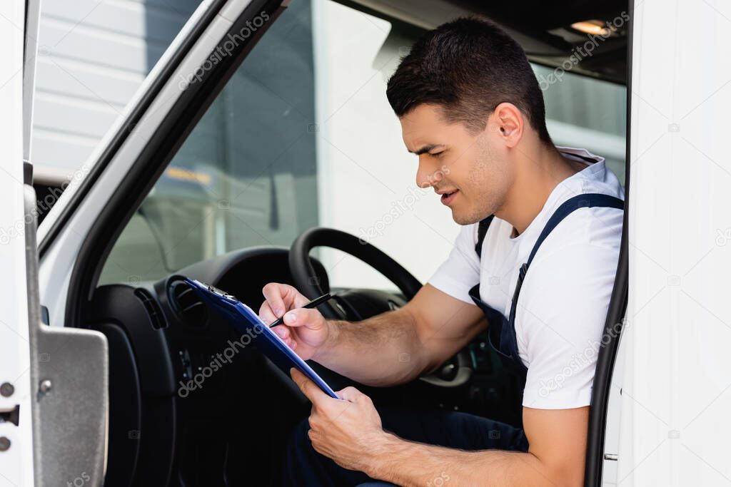 Selective focus of concentrated loader writing on clipboard while sitting on driver seat in truck 