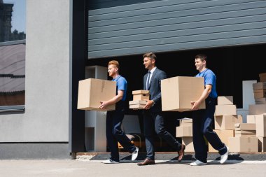 Businessman holding carton boxes while walking with movers on urban street  clipart
