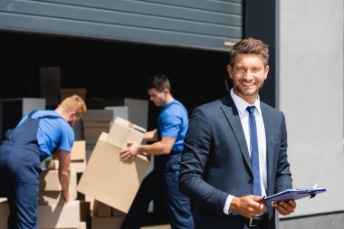 Selective focus of businessman with clipboard looking at camera while movers working in warehouse outdoors  clipart