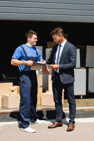 Loader holding carton box near man in suit writing on clipboard and warehouse at background