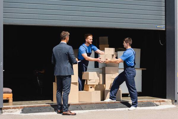 Businessman with clipboard standing near movers holding cardboard boxes in warehouse outdoors 