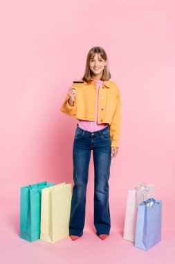 Pembe arka planda renkli alışveriş torbalarının yanında kredi kartı gösteren genç bir kadın.