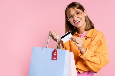 Güneş gözlüklü genç bir kadın elinde kredi kartı ve alışveriş torbalarıyla pembe arka planda fiyat etiketi var.