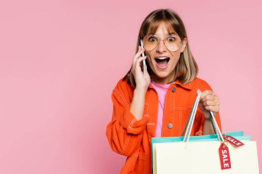 Shocked woman looking at camera while holding purchases with sale word on price tags and talking on smartphone on pink background clipart