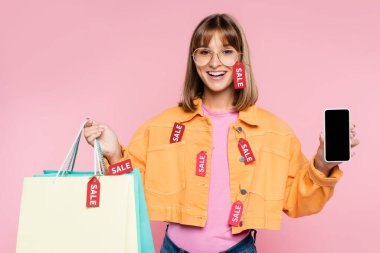 Young woman holding shopping bags with price tags and sale lettering and smartphone with blank screen on pink background clipart