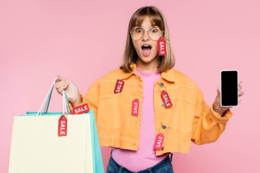 Shocked woman in sunglasses and price tags on jacket showing smartphone and colorful shopping bags on pink background clipart