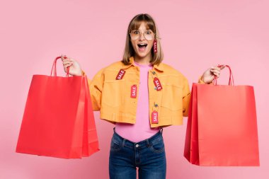 Sarı cekette fiyat etiketi olan şoke olmuş bir kadın pembe arka planda kırmızı alışveriş torbaları taşıyor.