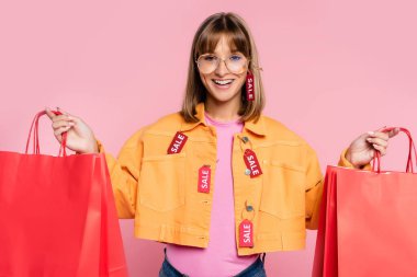 Ceketli ve güneş gözlüklü genç bir kadın. Pembe arka planda kırmızı alışveriş torbaları taşıyor.