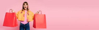Ceketinde fiyat etiketi ve güneş gözlüğü olan heyecanlı bir kadının pembe arka planda kırmızı alışveriş torbalarını tutarken kameraya bakışının yatay görüntüsü.