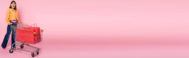 Pembe arka planda kırmızı alışveriş çantalarının yanındaki kameraya bakan heyecanlı kadının panoramik görüntüsü.