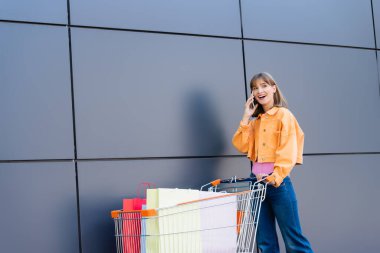 Cep telefonuyla konuşan bir kadın alışveriş çantalarının yanında, el arabasında ve bina cephesinde. 