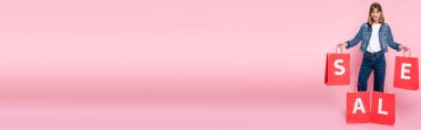 Pembe arka planda kırmızı alışveriş torbaları taşıyan panoramik genç bir kadın.