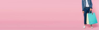 Pembe arka planda renkli alışveriş torbaları taşıyan kot pantolonlu bir kadın.