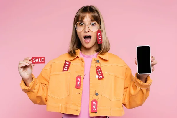 Surprised woman holding price tag with sale word and smartphone with blank screen on pink background