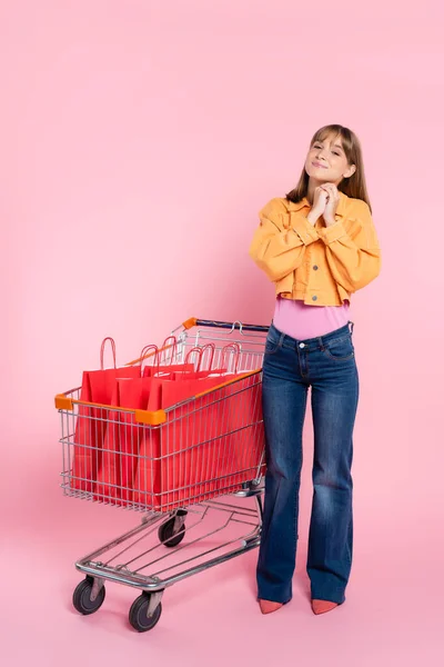 Junge Frau Blickt Kamera Neben Einkaufswagen Mit Roten Einkaufstüten Auf — Stockfoto