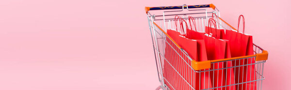 Panoramic shot of cart with red shopping bags on pink background