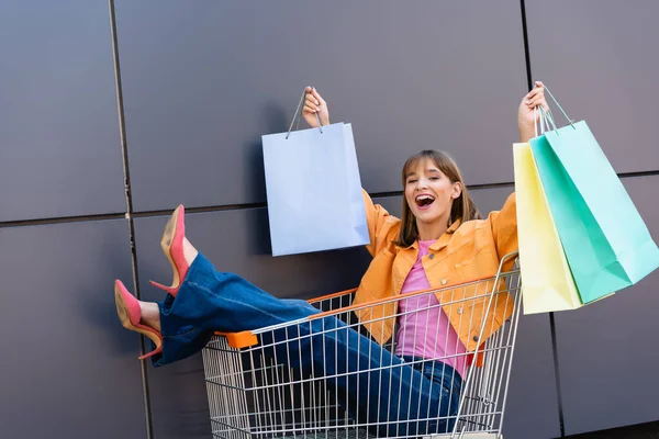 Aufgeregte Frau Sitzt Einkaufswagen Und Hält Einkaufstüten Der Nähe Des — Stockfoto