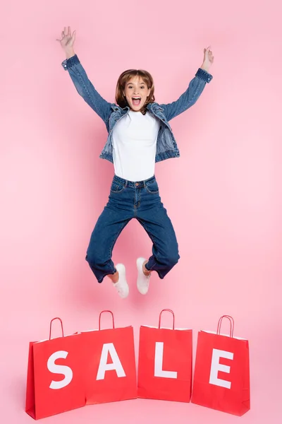 Aufgeregte Frau Springt Neben Einkaufstüten Mit Verkaufswort Auf Rosa Hintergrund — Stockfoto