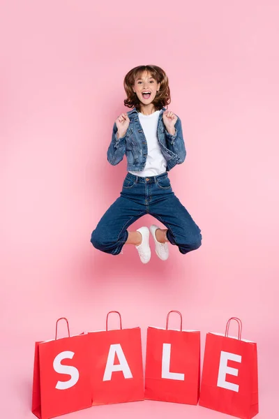 Excited Woman Jeans Jumping Shopping Bags Sale Lettering Pink Background — Stock Photo, Image
