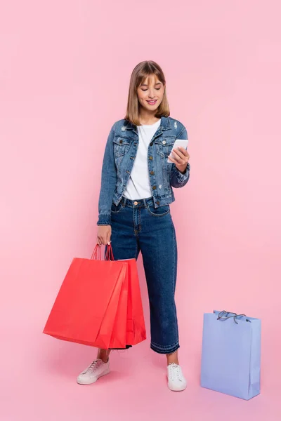 Mujer Joven Usando Smartphone Sosteniendo Bolsas Compras Sobre Fondo Rosa — Foto de Stock