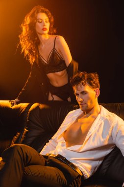 Takım elbiseli adam kanepede oturuyor. Dantelli iç çamaşırlı, siyah ceketli baştan çıkarıcı bir kadının yanında. 