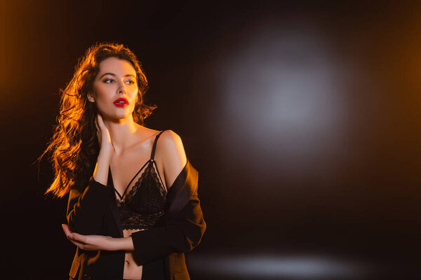 соблазнительная и кудрявая женщина в кружевном бюстгальтере и блейзере смотрит в сторону черного 