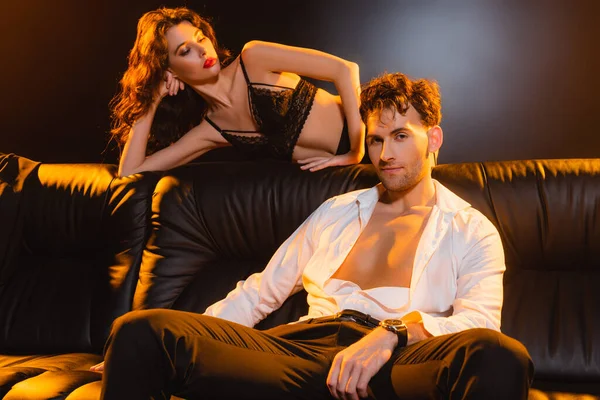 man in suit sitting on sofa and looking at camera near seductive woman in lace underwear on black
