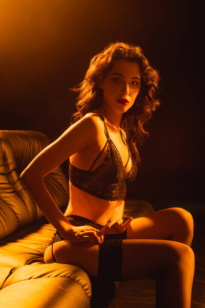 кудрявая молодая женщина в кружевном белье сидит на диване на черном