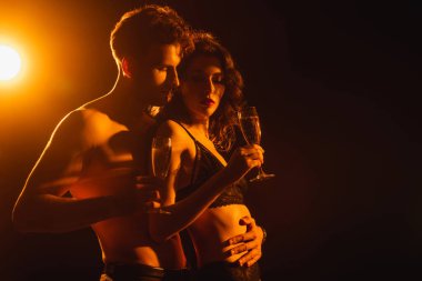 Üstsüz bir adam, baştan çıkarıcı kız arkadaşına sarılıyor. Siyah üzerine bir kadeh şampanya ve arka ışık.