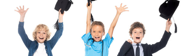 panoramic shot of excited kids dressed in costumes of different professions holding graduation caps above heads isolated on white 