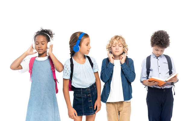 schoolgirl listening music in wireless headphones and looking at curly boy reading book near multicultural schoolchildren isolated on white 