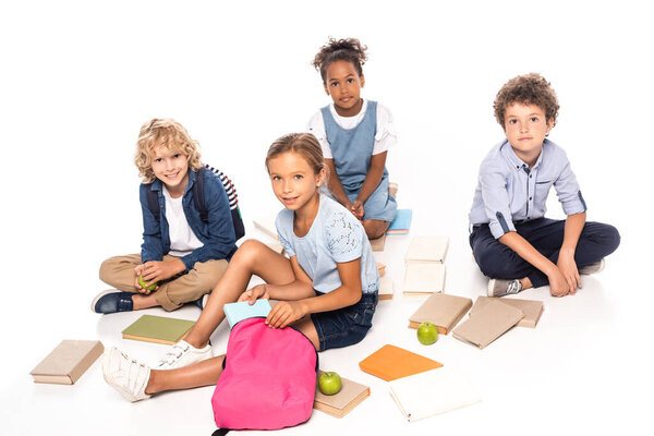 multicultural schoolkids sitting near books and apples isolated on white