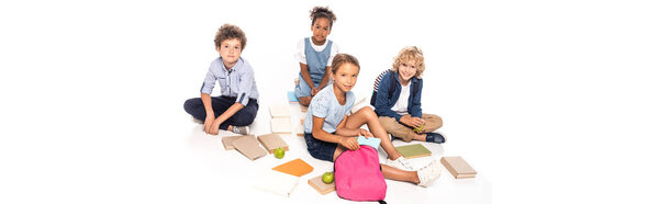 panoramic crop of schoolkids sitting near books, apples and african american kid in wireless headphones isolated on white