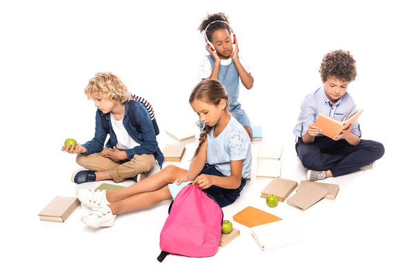 schoolchildren sitting near books, apples and african american kid in wireless headphones isolated on white
