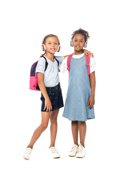schoolgirl listening music in wireless headphones and hugging african american friend isolated on white 
