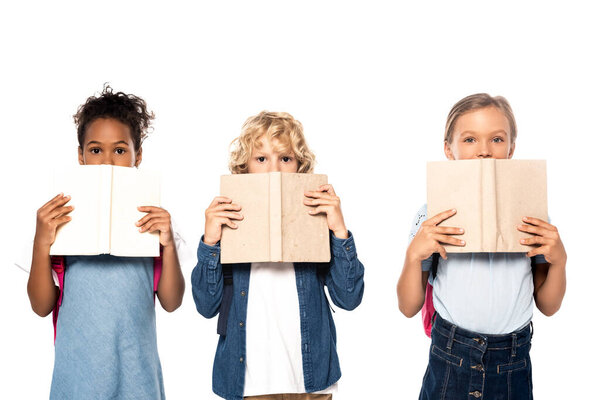 multicultural schoolgirls and schoolboy covering faces with books and looking at camera isolated on white 
