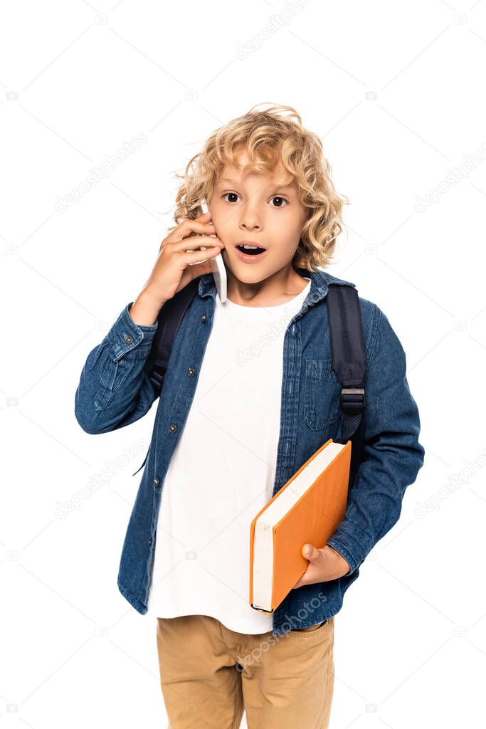 shocked and blonde schoolboy holding book and talking on smartphone isolated on white 