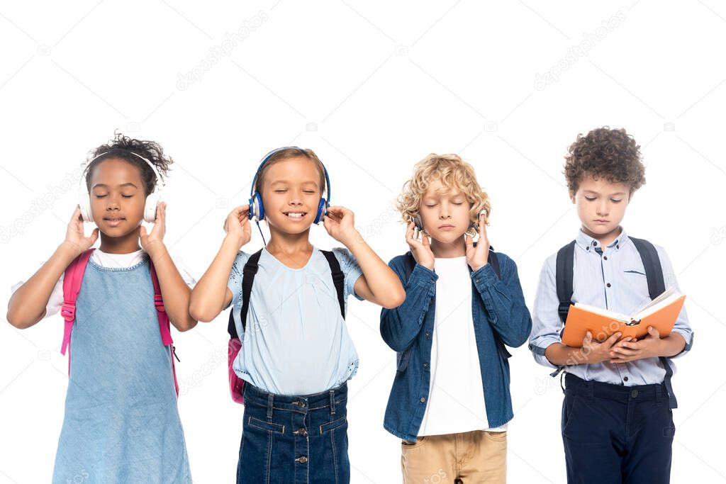 multicultural schoolchildren listening music in wireless headphones near curly boy reading book isolated on white 