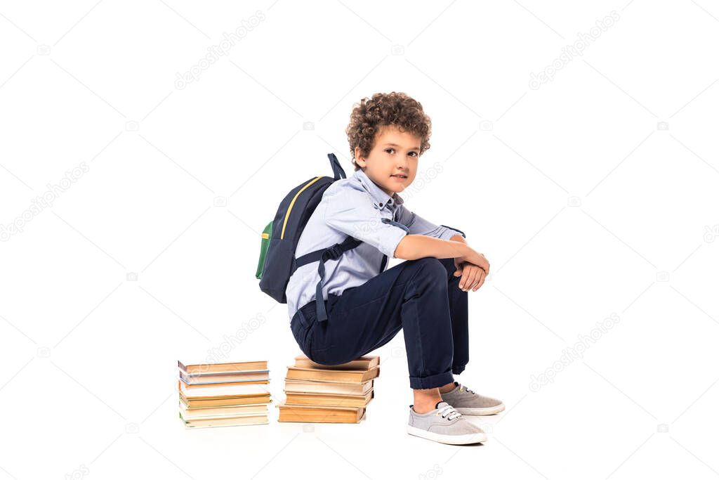 curly schoolboy with backpack sitting on books isolated on white 