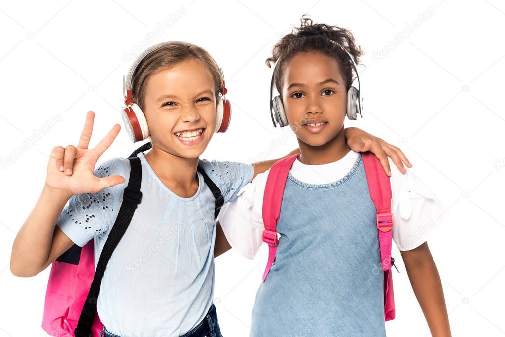 schoolkid listening music in wireless headphones, hugging african american friend and showing three fingers isolated on white 
