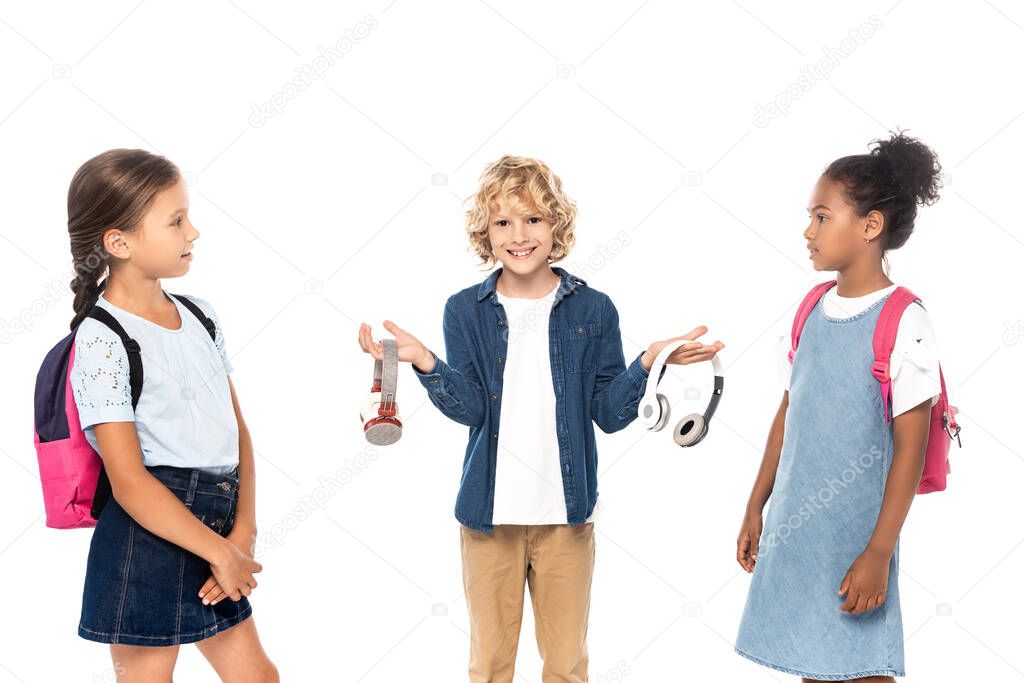 blonde schoolboy holding wireless headphones near multicultural schoolgirls isolated on white 